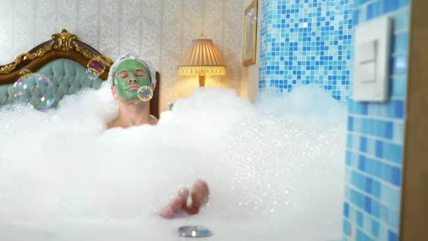 Homem bonito emocional com máscara de barro em uma touca de banho soprando bolhas de sabão deitado em uma banheira com espuma abundante em um banheiro luxuoso. espaço de cópia — Vídeo de Stock