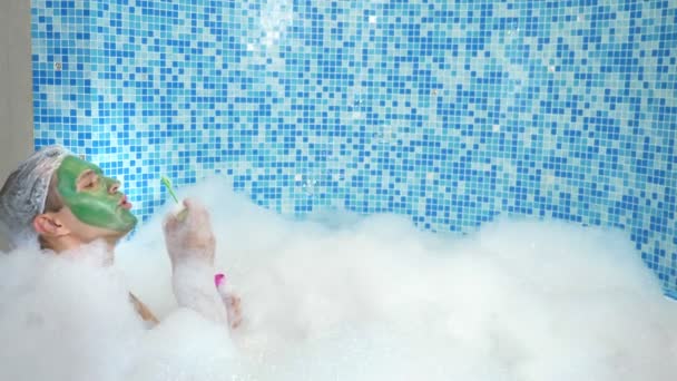 Hombre lindo emocional con máscara de arcilla en una gorra de baño, soplando burbujas de jabón mientras está acostado en una bañera con abundante espuma. concepto humorístico. espacio de copia — Vídeo de stock