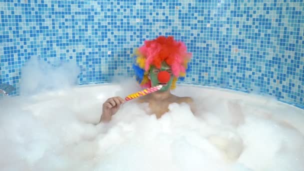 泡の多い浴槽で可愛いピエロさんが棒にロリポップを食べます。ユーモアのあるコンセプト. — ストック動画