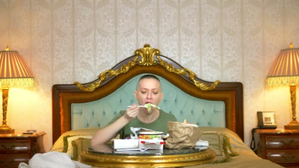 Πεινασμένο φαλακρό κορίτσι σε ένα χακί πουκάμισο τρώει fast food κάθεται σε ένα πολυτελές εσωτερικό στο κρεβάτι — Αρχείο Βίντεο