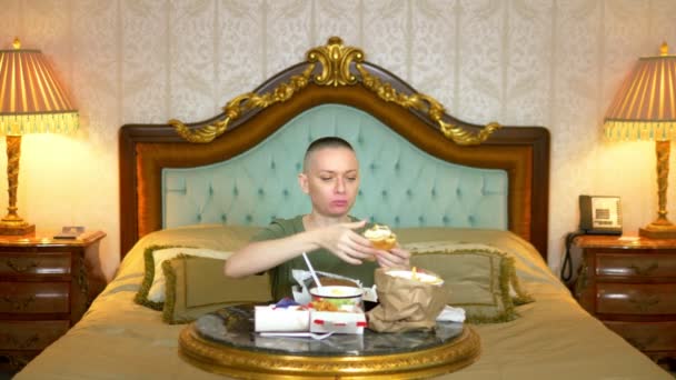 Careca faminto menina em uma camisa cáqui comer fast food sentado em um interior luxuoso na cama — Vídeo de Stock