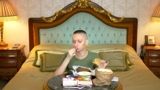 Chica calva hambrienta en una camisa caqui comiendo comida rápida sentado en un interior de lujo en la cama — Vídeo de stock