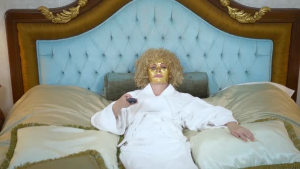 Junge blonde Frau mit goldener Maske im Gesicht vor dem Fernseher auf einem luxuriösen goldenen Bett liegend. — Stockvideo