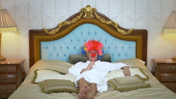 Un jeune homme en manteau blanc et une perruque de clown gît sur un lit doré luxueux et mange une sucette sur un bâton. concept d'humour, aventure de personnes étranges . — Video