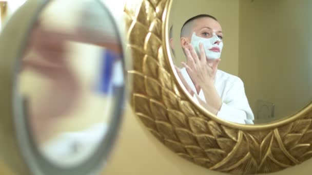 Chica calva en un abrigo blanco pone una máscara de belleza en su cara, mirando en el espejo en el baño . — Vídeo de stock