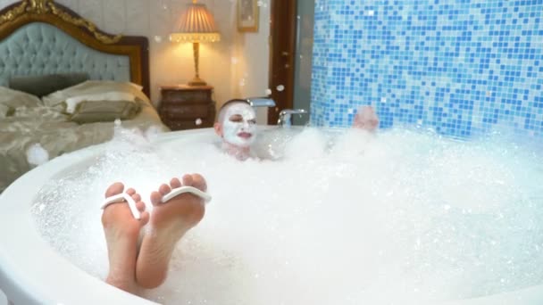 Eine glatzköpfige junge Frau mit Maske im Gesicht liegt in einem Schaumbad, ihre Beine mit Zehentrenner sind vom Wasser aus sichtbar. — Stockvideo