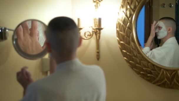 Chauve fille dans un manteau blanc met un masque de beauté sur son visage, regarder dans le miroir dans la salle de bain . — Video