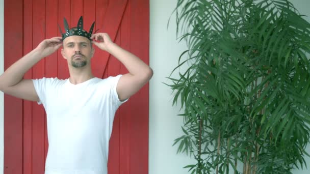 Pohledný podivný muž v bílém tričku klade na hlavu koženou černou korunu — Stock video