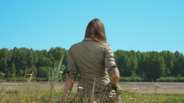Vista trasera, chica de pelo justo con el pelo largo se sienta en la orilla del río — Vídeos de Stock