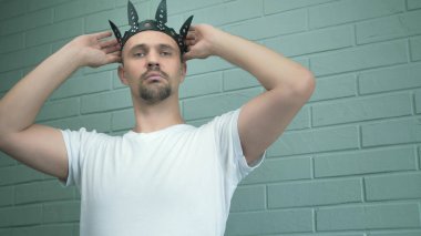 Beyaz tişörtlü yakışıklı adam başına siyah deri bir taç takıyor.