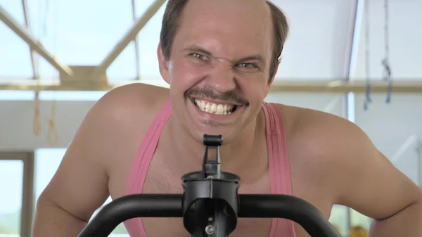 Közelről, vicces kopasz férfi edzés egy torna kerékpár, mosolygós — Stock Fotó