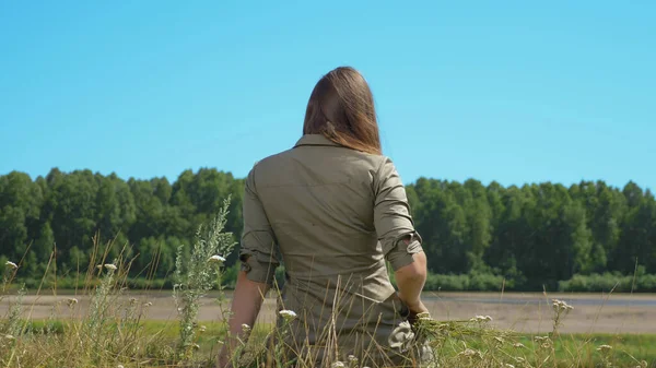 Vista trasera, chica de pelo justo con el pelo largo se sienta en la orilla del río —  Fotos de Stock