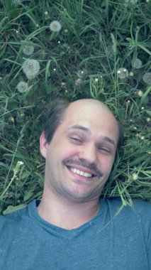 Dikey olarak. Genç ve kel bir adam çimlerin üzerinde uzanmış karahindiba ve gülümsemeyle