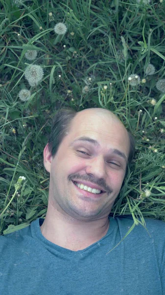 vertically. a young balding man lying on the grass with dandelions and smiles