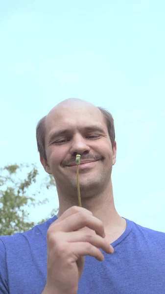 Vertically. young balding man blows on a dandelion on a background of blue sky — Stock Photo, Image