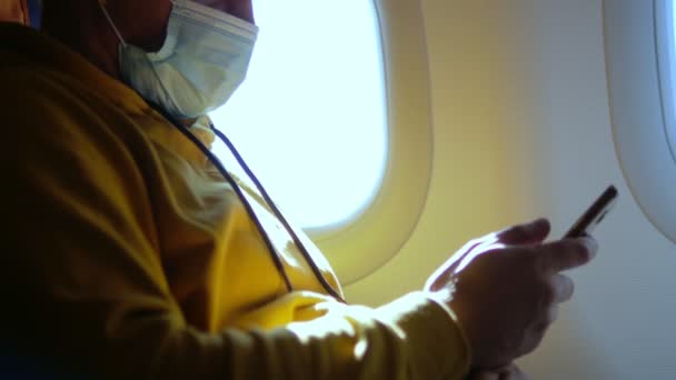 Närbild av en man i medicinsk mask med hjälp av en smartphone på ett flygplan — Stockvideo