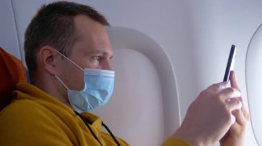 Uçakta akıllı telefon kullanan tıbbi maskeli bir adamın yakın plan fotoğrafı.