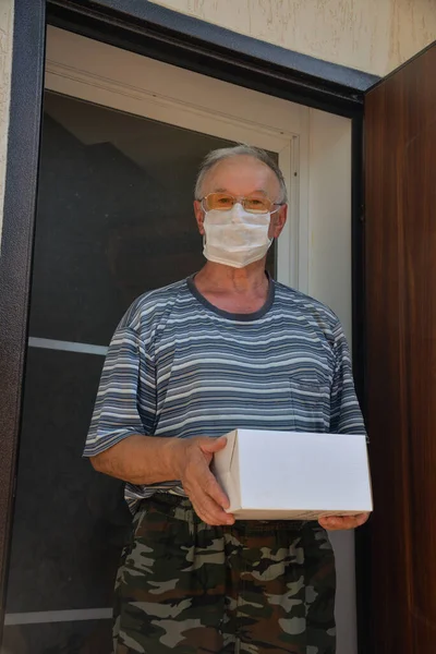 Koruyucu maskeli bir adam elinde teslimat servisi tarafından alınmış bir kutuyla bahçede duruyor.