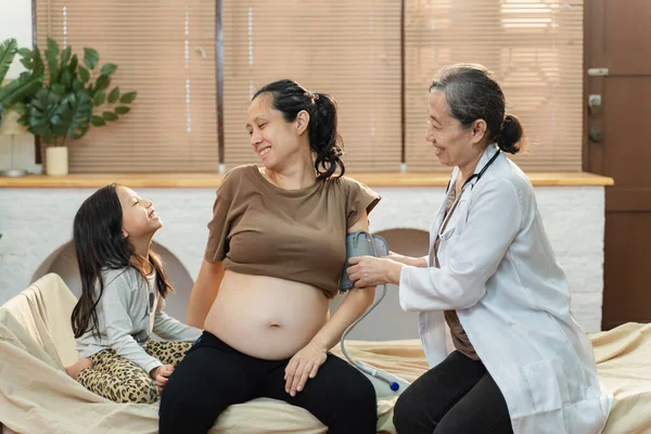 Genç Asyalı hamile kadın ve çocukla ev ziyareti sırasında hemşire. Başhekim hastanın tansiyonunu ölçüyor.