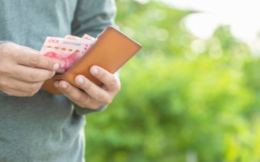 Asyalı adam cüzdanındaki Çin banknotlarını sayıyor ve kontrol ediyor. Yeşil bulanık arkaplan ile açık havada çekim