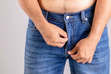 Gri arka planda kot pantolonunun fermuarını çekmeye çalışan şişman adamı kapat.
