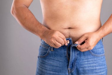 Gri arka planda pantolon stüdyosunun düğmelerini iliklemeye çalışan şişman adamı kapat.