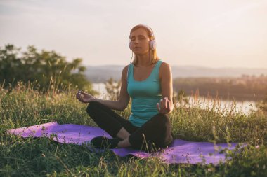 Güzel sportif ve kadın ile heaphones meditasyon egzersiz üzerinde otururken müzik dinleme ile cityscape ve nehir behinde onu mat. Görüntü kasten tonda.
