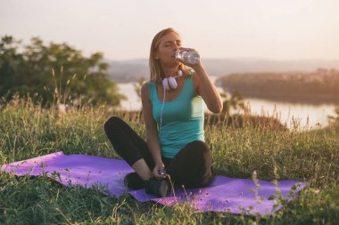 Egzersiz üzerinde otururken güzel sportif kadın içme suyu ile cityscape ve nehir behinde onu mat. Görüntü kasten tonda.