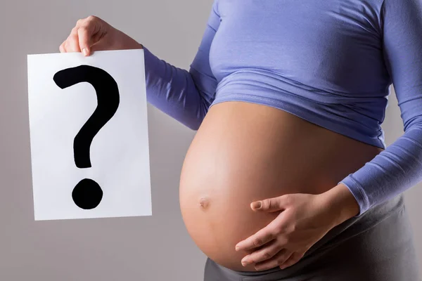 灰色の背景に疑問符が付いた紙を持つ妊婦のクローズアップ胃の画像 — ストック写真