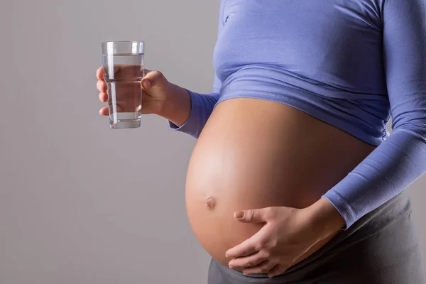 灰色の背景に水のガラスを保持している妊婦のクローズアップ胃の画像 — ストック写真