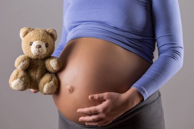 Gri arka planda oyuncak ayı tutan hamile kadının yakın mide görüntüsü.