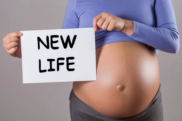 灰色の背景にテキスト新しい生活と紙を保持している妊娠中の女性のクローズアップ胃の画像 — ストック写真