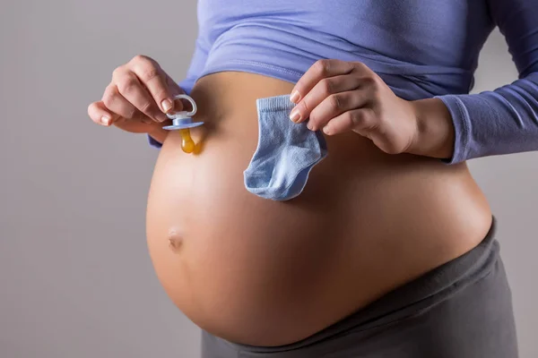 在灰色背景上为男婴拿着袜子和奶嘴的孕妇胃图 — 图库照片