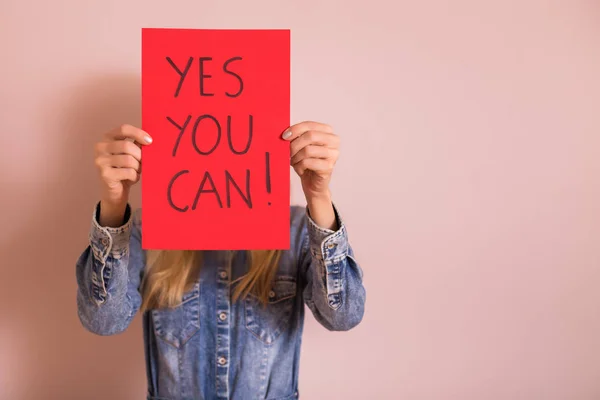 Woman holding paper with text yes you can while standing in front of the wall.