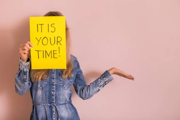 Woman holding paper with text it is your time while standing in front of the wall.