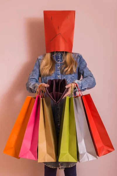 Sad woman holding shopping bags and empty wallet while standing in front of the wall.