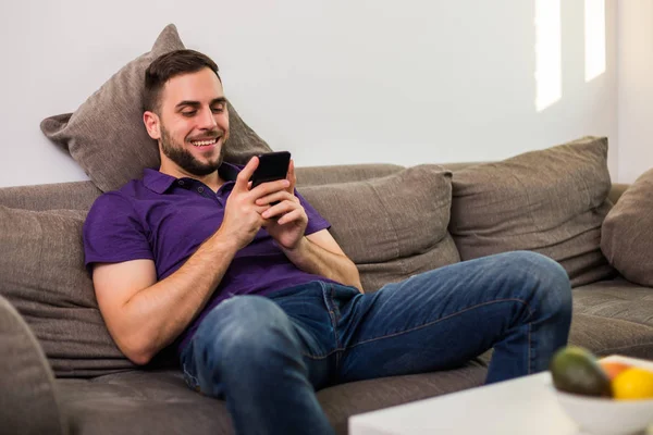 リビングルームのソファに座りながら携帯電話を使う若い男 — ストック写真