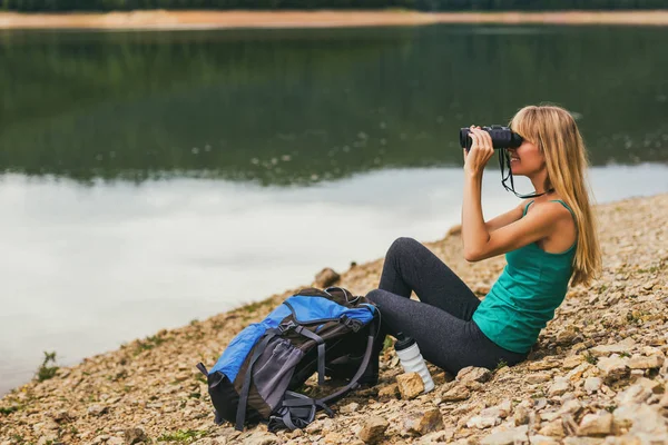 湖のそばに座っている間双眼鏡を使った女性ハイカー — ストック写真