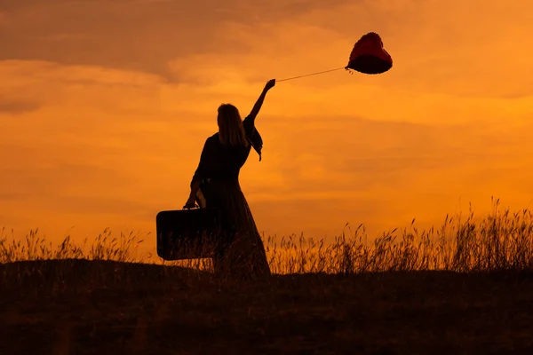 美しい夕日を眺めながら スーツケースとハート型の風船を持つ女性のシルエット — ストック写真