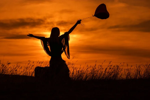 ハート型の風船でスーツケースに座って美しい夕日を見ている女性のシルエット — ストック写真