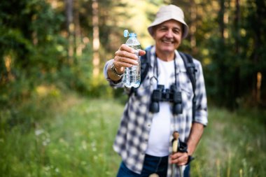 Portrait of happy senior man drinking water while enjoying hiking with backpack and hiking poles in nature. Focus on water bottle.