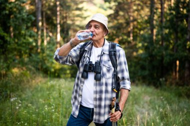 Portrait of happy senior man drinking water while enjoying hiking with backpack and hiking poles in nature.
