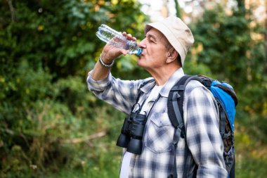 Doğa yürüyüşünün keyfini çıkarırken sırt çantasıyla içme suyu içen kaygısız yaşlı adamın portresi..