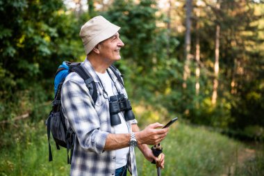Sırt çantası ve yürüyüş direkleri olan kaygısız yaşlı bir adamın portresi. Doğada yürüyüş yaparken cep telefonu kullanıyor..