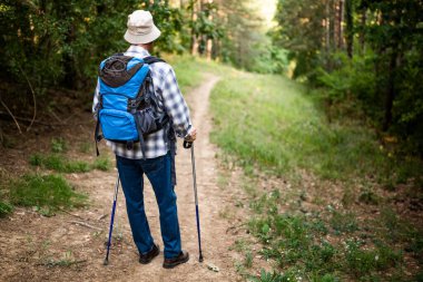 Senior man hiking along mountainous forest path with backpack and hiking poles. He is enjoying the freedom, relaxation and discovery that comes with outdoor exploration.