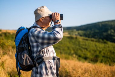 Doğa yürüyüşü sırasında dürbün kullanan sırt çantalı yaşlı bir adamın portresi.