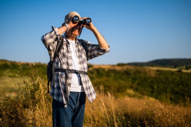Doğa yürüyüşü sırasında dürbün kullanan sırt çantalı yaşlı bir adamın portresi.