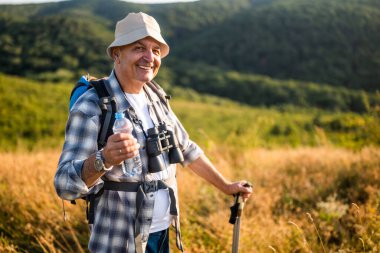 Portrait of happy senior man drinking water while enjoying hiking with backpack and hiking poles in nature.
