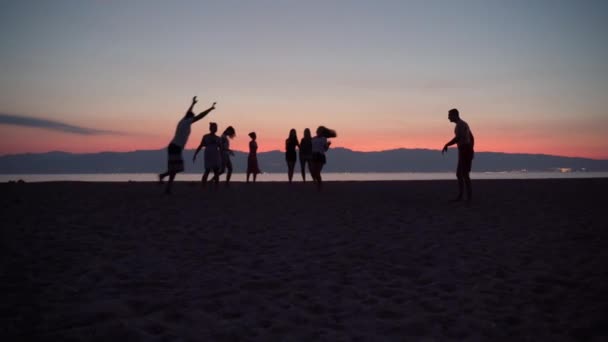 Güzel deniz günbatımı arka planında dans eden neşeli insanların siluetleri. Arkadaşlar dans ediyor ve zıplıyor, mutlu adam kumların üzerinde dans ediyor. Sıcak plaj partisi konsepti — Stok video