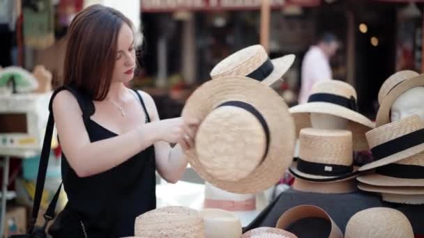 Mercado de artesanato incrível cheio de diferentes produtos artesanais, menina bonita com sorriso adorável tentando no chapéu de palha de verão elegante e olhando para o espelho. Escolha entre estilos de chapéus na pulga local — Vídeo de Stock